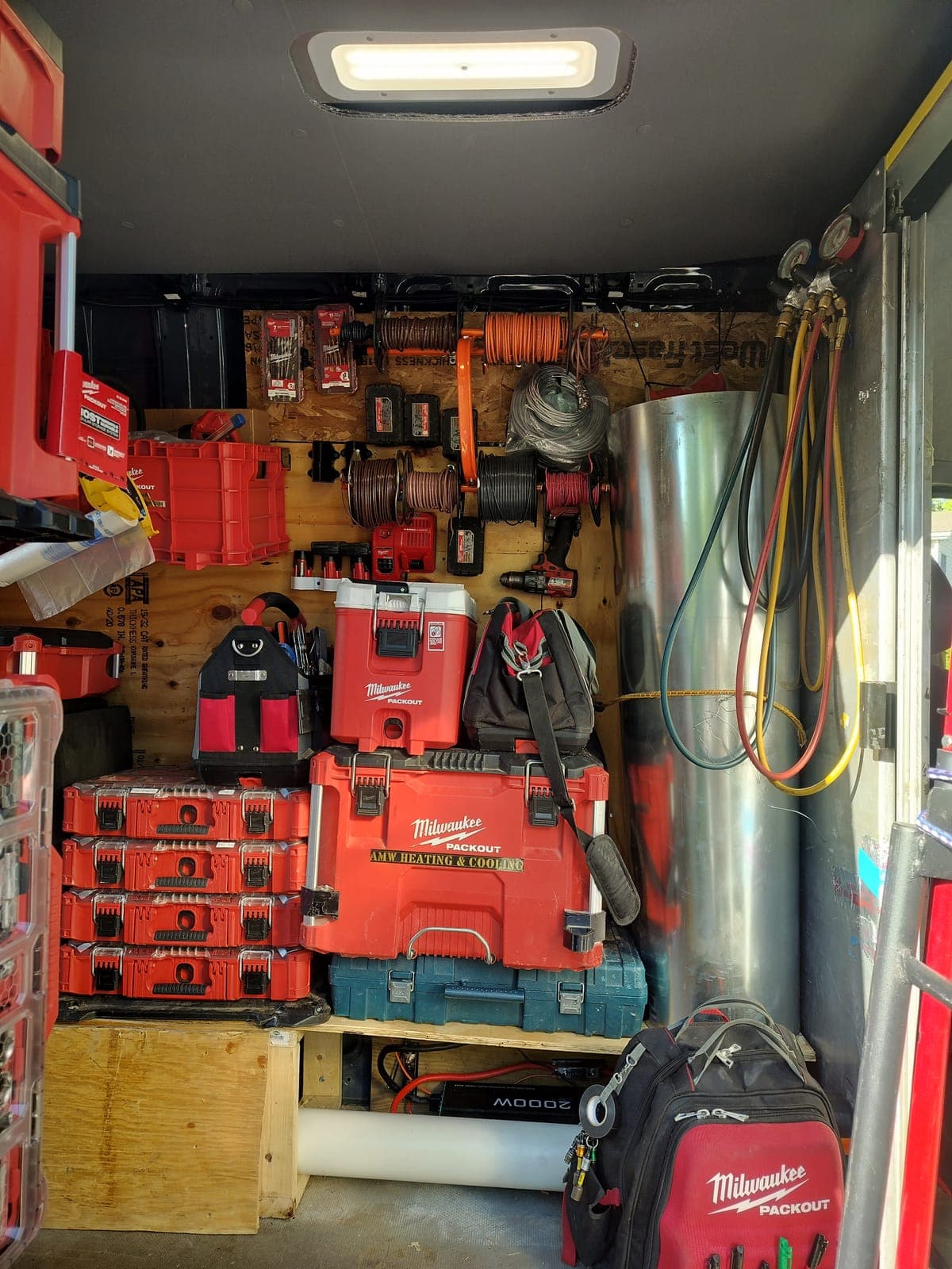 Inside an organized AMW Heating & Cooling service van, labeled Milwaukee Packout cases, hose reels, and refrigerant gauges mounted on the wall